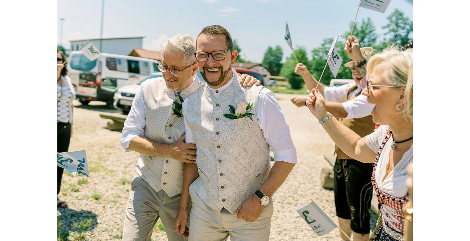 Zwei Männer nach der standesamtlichen Trauung auf einem Floß in Lechbruck am See, umgeben von Gästen in Tracht, die sie mit kleinen Fahnen begrüßen