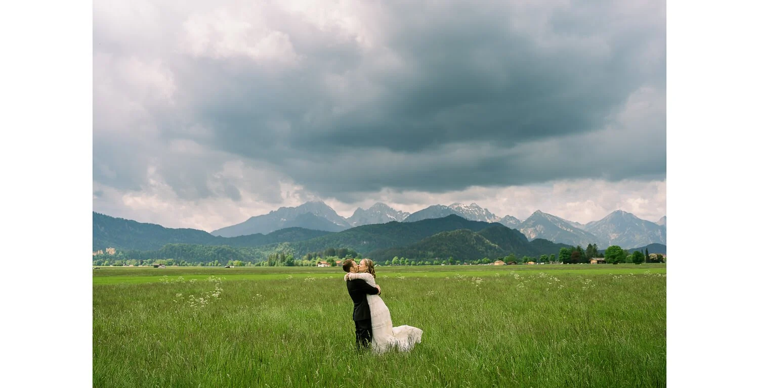 Paarshooting auf einer Wiese bei Füssen, Brautpaar umarmt sich vor weiter Alpenlandschaft und dramatischem Himmel