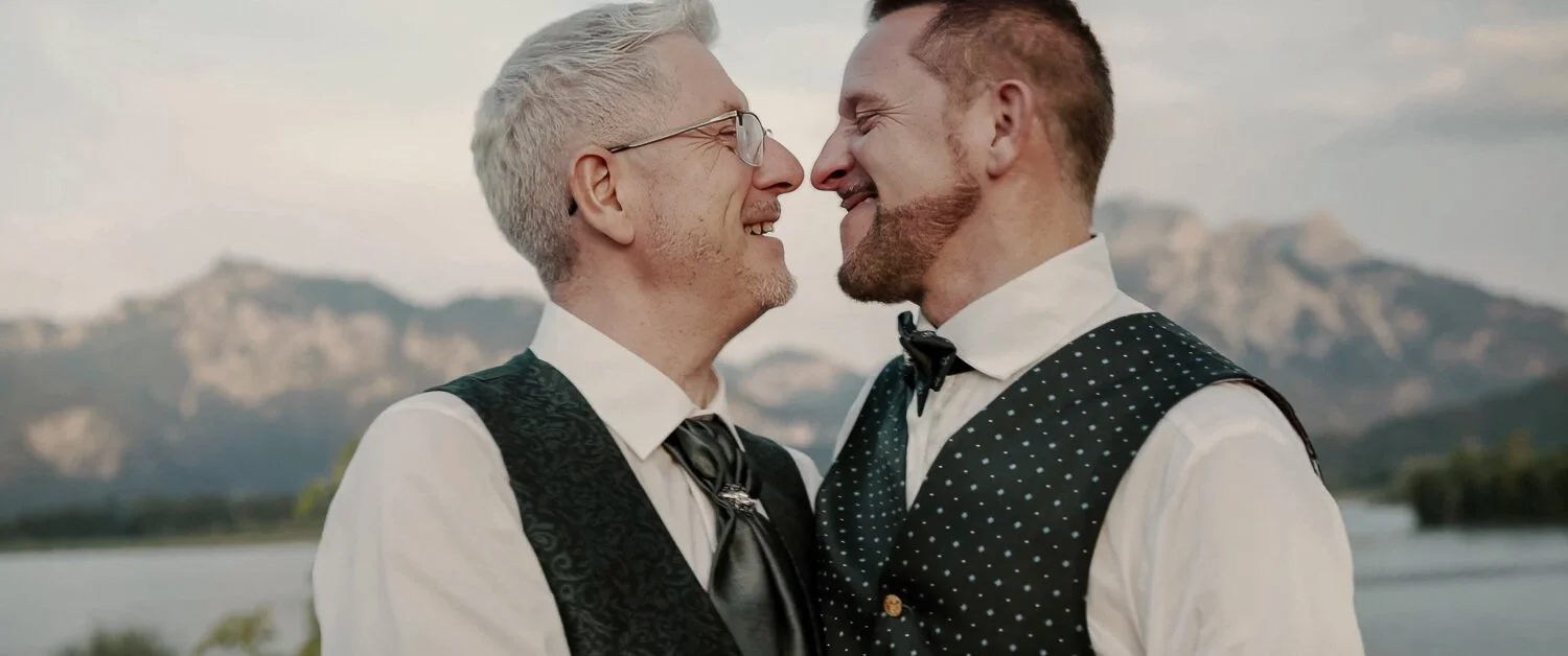 Zwei Männer stehen sich lächelnd gegenüber, im Hintergrund der Forggensee und die Berge bei Füssen, aufgenommen während ihrer Hochzeit.