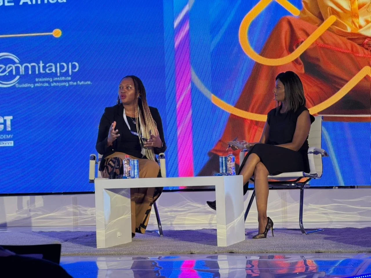 Mimi Kalinda and Alishia Seckam participating in a panel discussion on stage with a large blue screen behind them. Mimi is speaking, while the other is listening and taking notes, both seated on chairs with small tables in front of them.