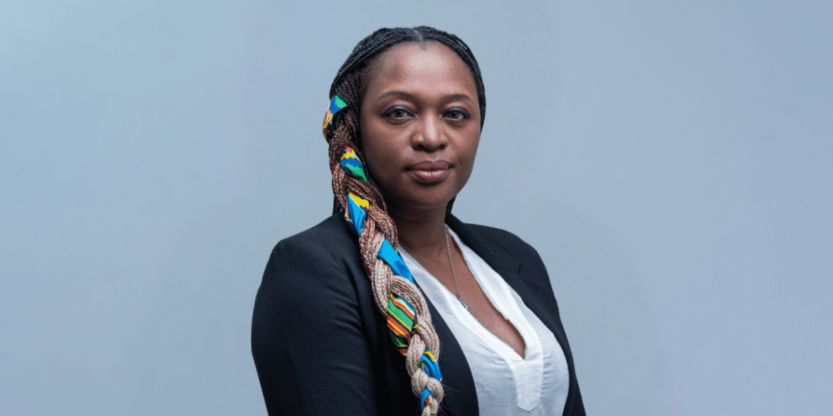 Mimi Kalinda with braided hair decorated with colorful beads, wearing a black blazer over a white blouse, standing against a gray background for CEO Times article.