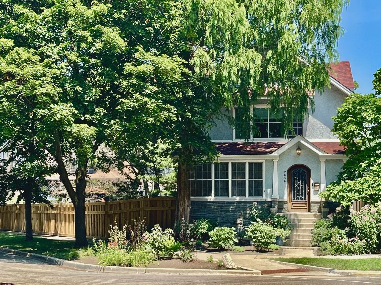 A two-story house with a white exterior, dark window frames, and a red front door, surrounded by green trees and bushes, with a wooden fence on one side and a small garden in front, on a sunny day in Chicago's Ravenswood Gardens neighborhood.