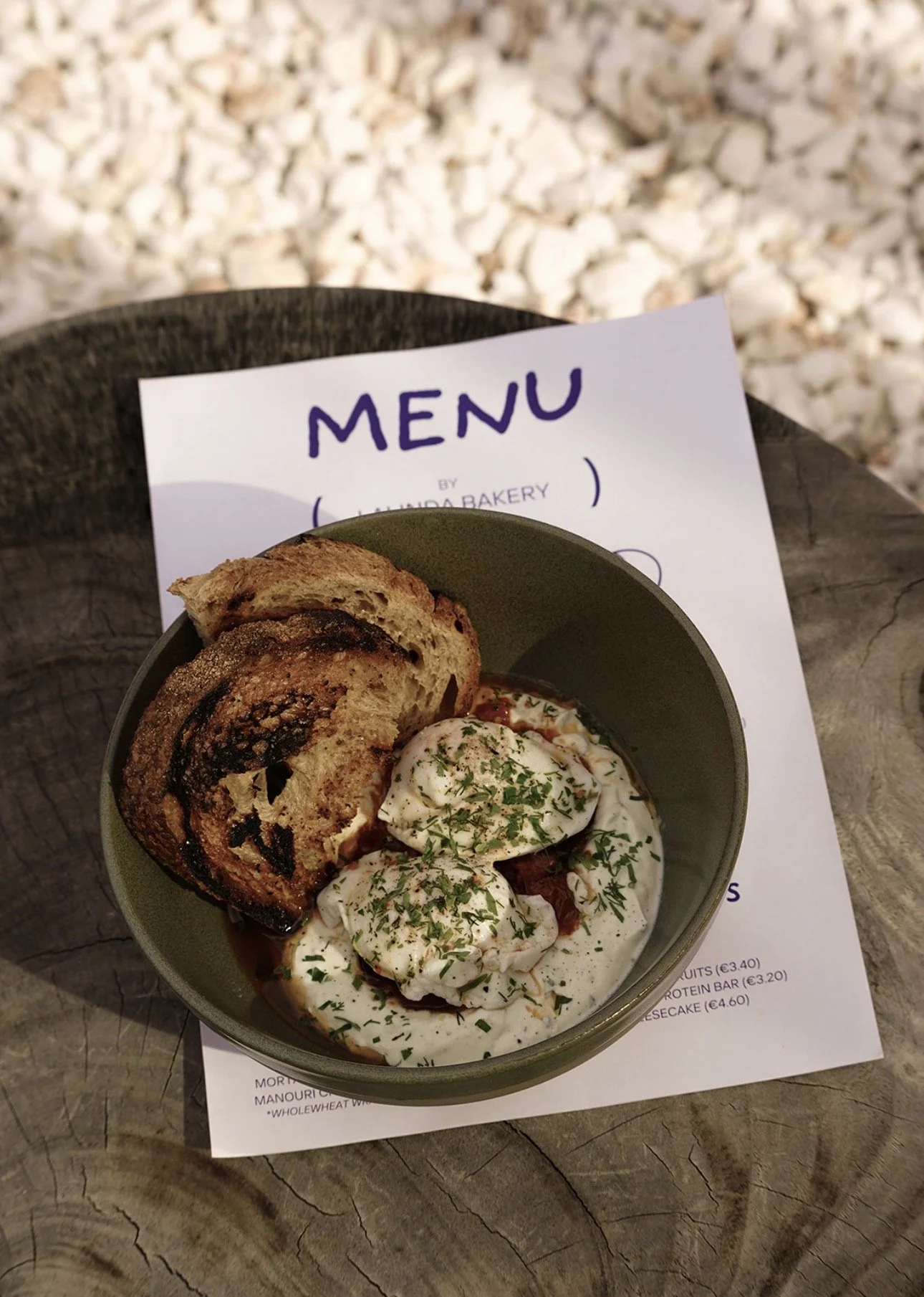 A bowl of poached eggs with creamy white sauce and chopped herbs, served with a slice of swirled bread on a wooden table, with a menu in the background.