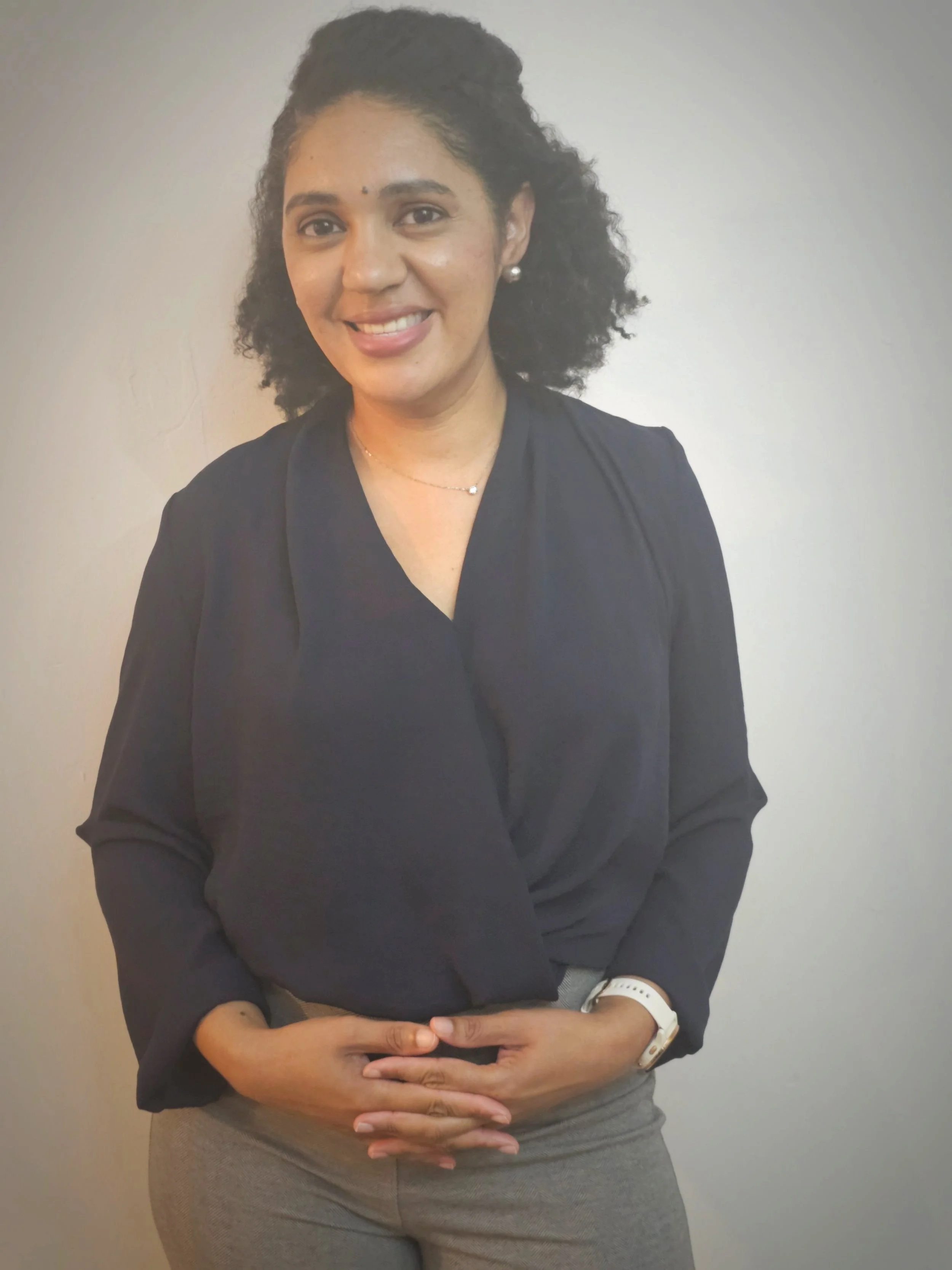 Person wearing a dark blouse and gray pants, smiling with hands clasped in front of them against a plain background.