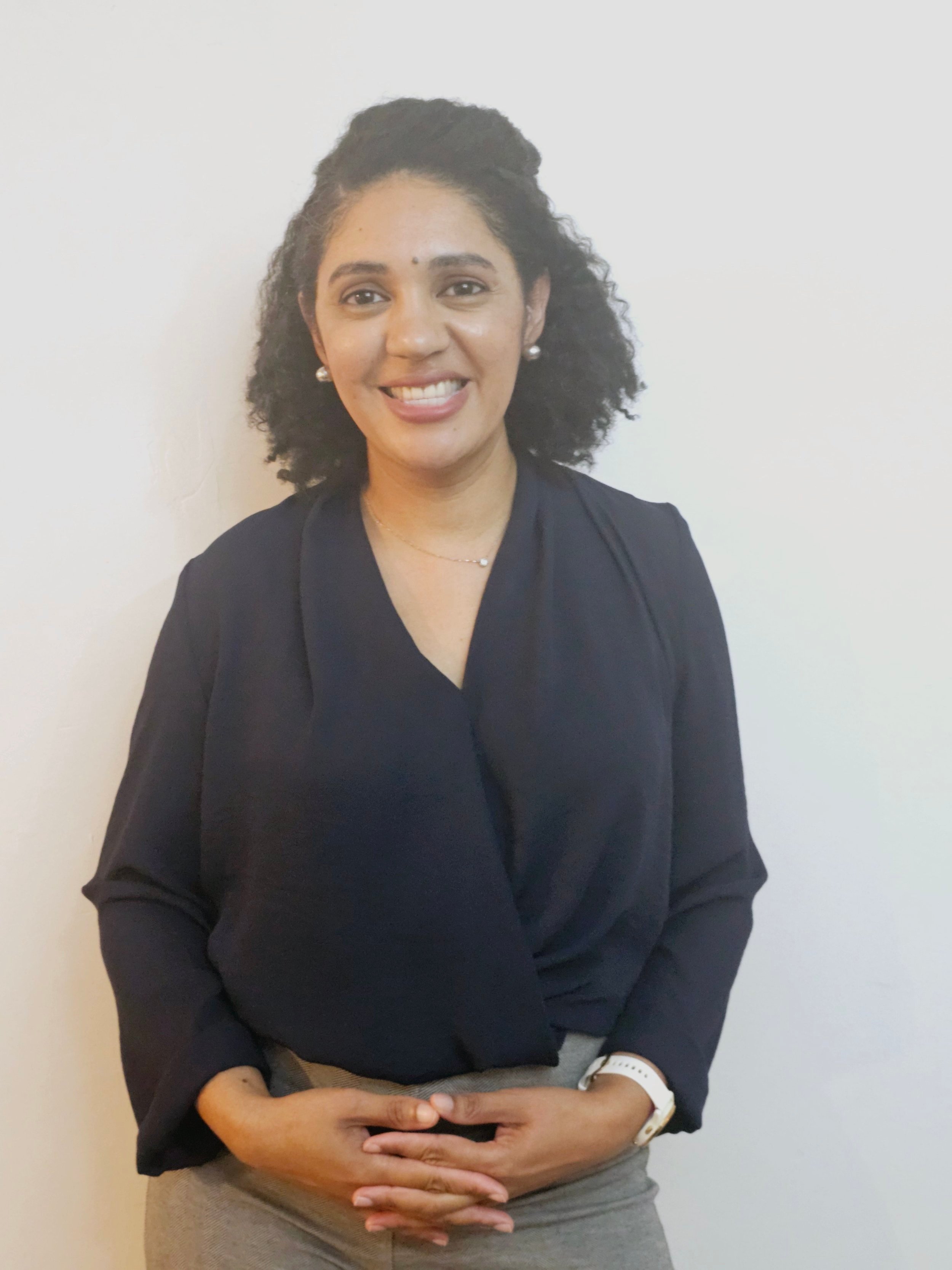 Smiling person with curly hair wearing a dark top, standing against a light background.