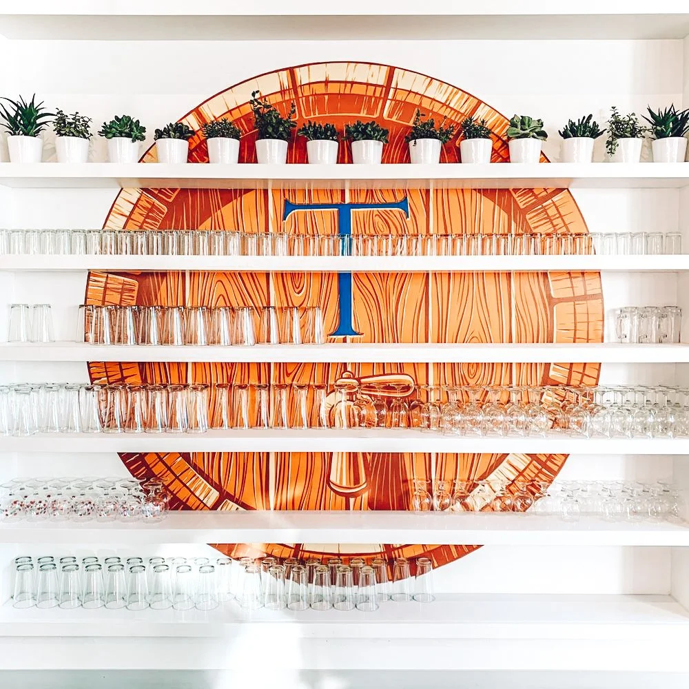 White shelves with potted plants, glasses, and decorative items in front of a large orange clock with a blue letter T in the center.