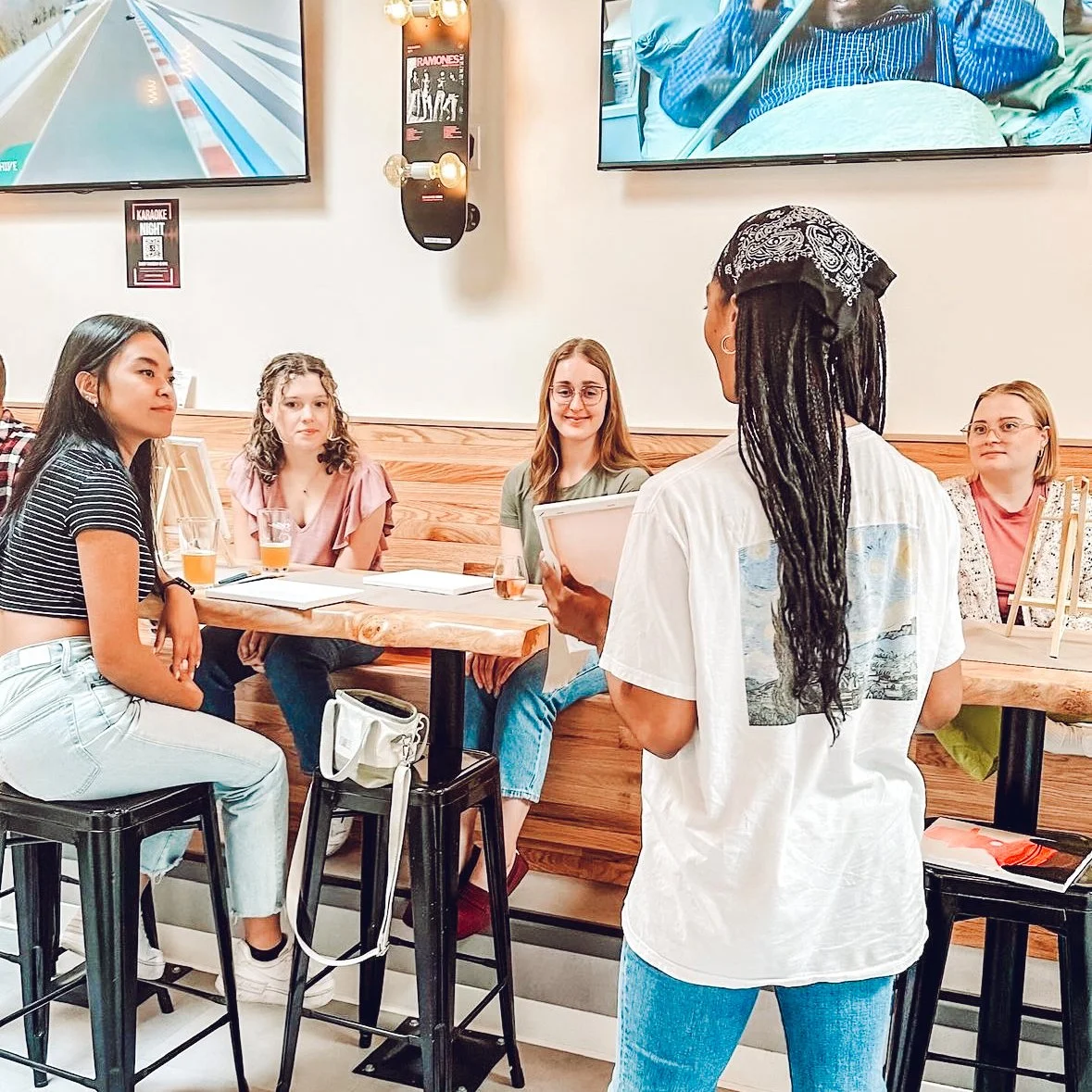 A group of four women sitting at a wooden table in a Tapster, listening to a woman with long dreadlocks and a bandana, who is reading from a sheet of paper. There are drinks on the table and two large TV screens on the wall.
