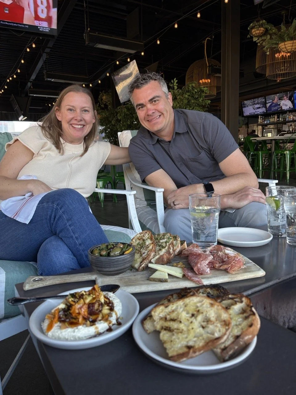 A smiling woman and man sitting at a table in a restaurant with drinks and a spread of food including pizza, cheese, cured meats, olives, and a salad.