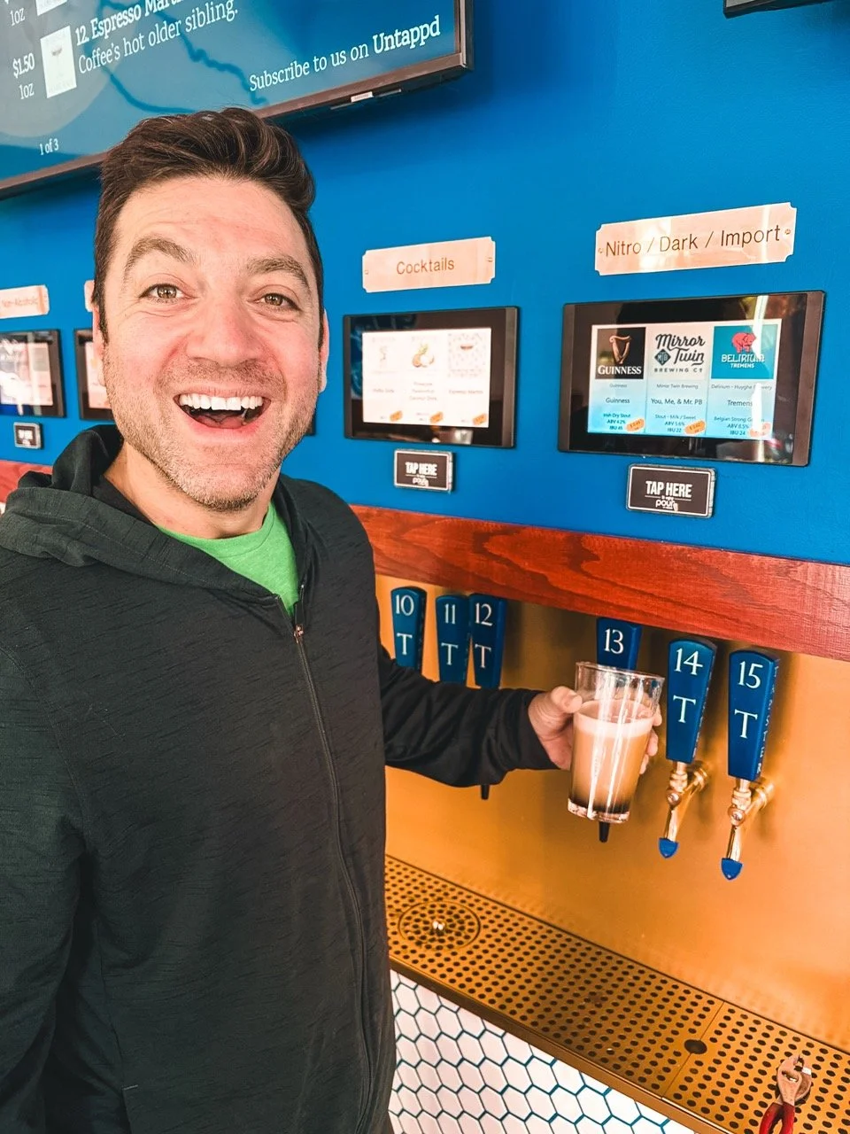 A man with short brown hair and a beard smiling happily while holding a glass of beer at a self-serve tap station in a brewery or bar. He is wearing a black hoodie and a green shirt underneath. The background includes a blue wall with menu screens and signs for drinks, with taps labeled 10 to 15.