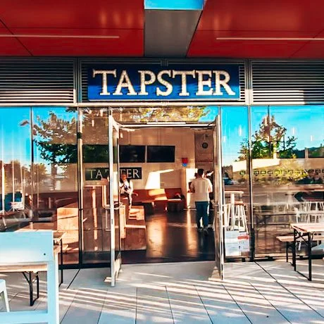 Front entrance of the Tapster in Seattle (South Lake Union) with outdoor seating area, glass doors, and a blue sign with white lettering.