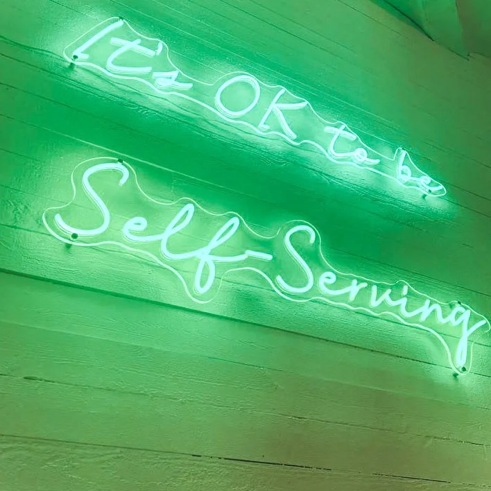 A neon sign with green glow that reads 'It's OK to be Self-Serving' on a wooden wall.