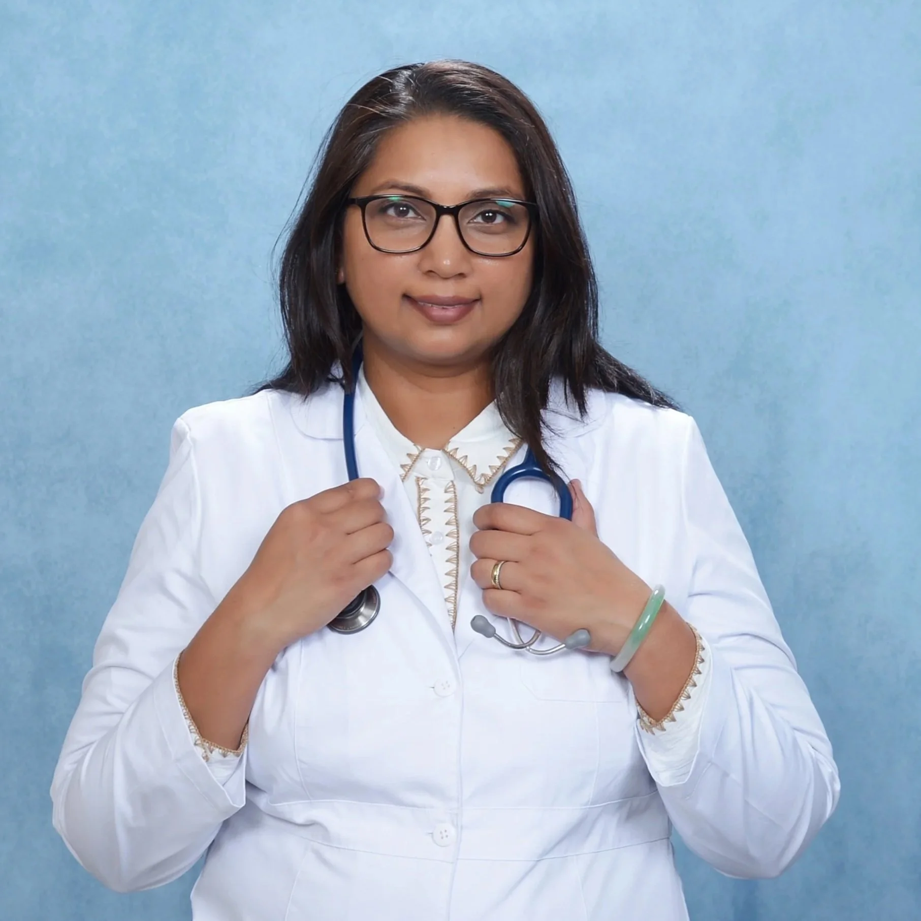 A woman doctor wearing glasses, a white coat, and a stethoscope around her neck, standing against a blue background.