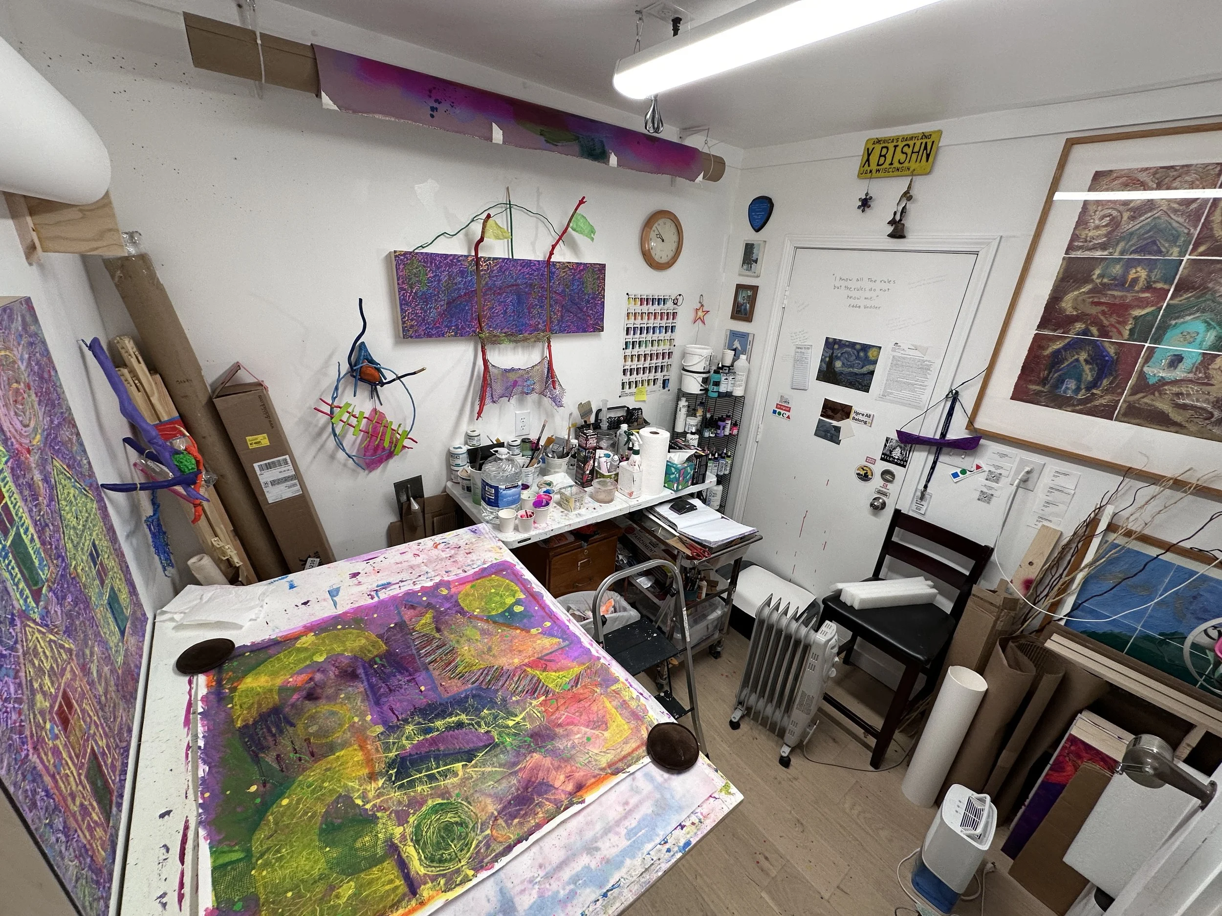 The artist’s intimate studio with paintings, supplies, and colorful art pieces on the walls and work table.