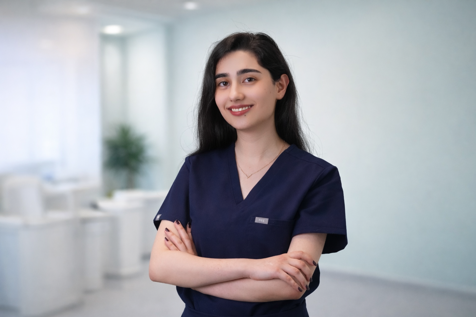 A young woman with long black hair wearing navy scrubs standing with arms crossed in a professional setting.