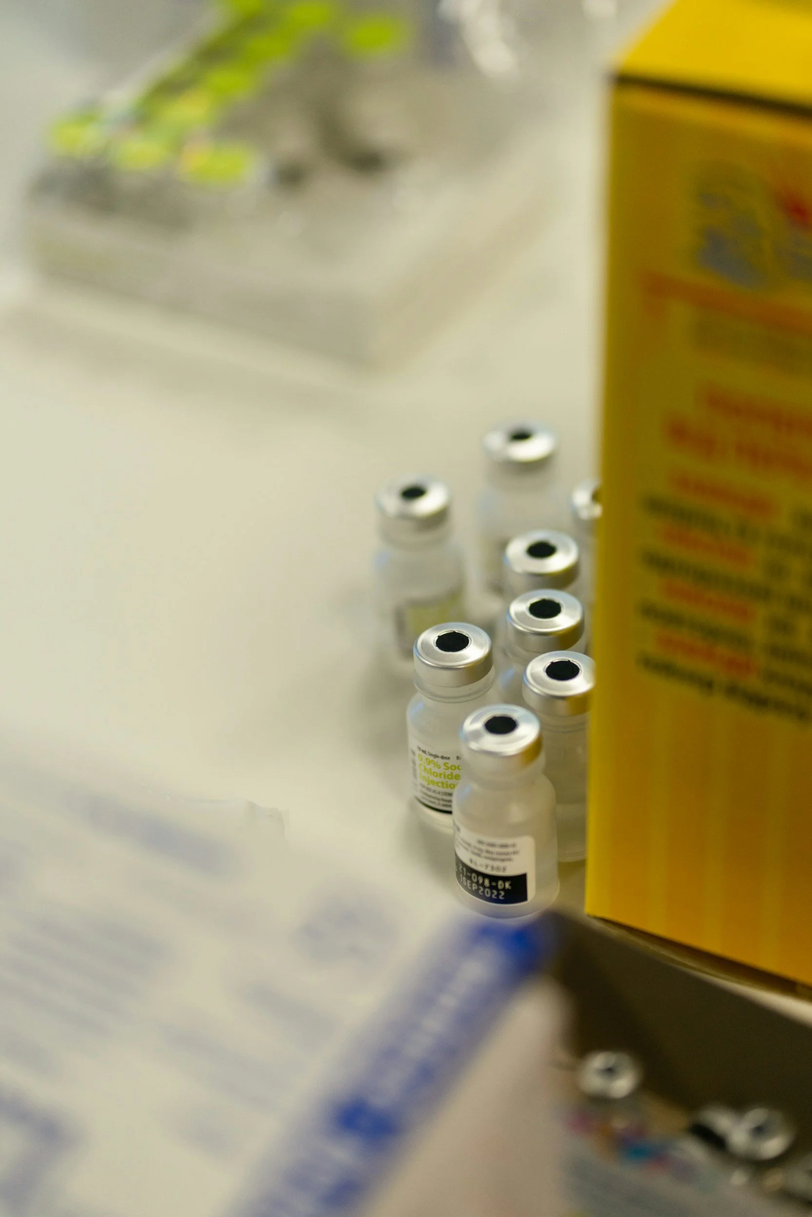 Several small glass vials with silver caps and black tops, arranged in a row on a white surface. A partially visible yellow box is on the right side. Blurry object in the background.
