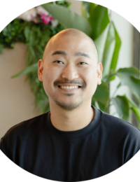 Smiling Asian man with a shaved head wearing a black shirt, standing indoors near green plants and pink flowers.