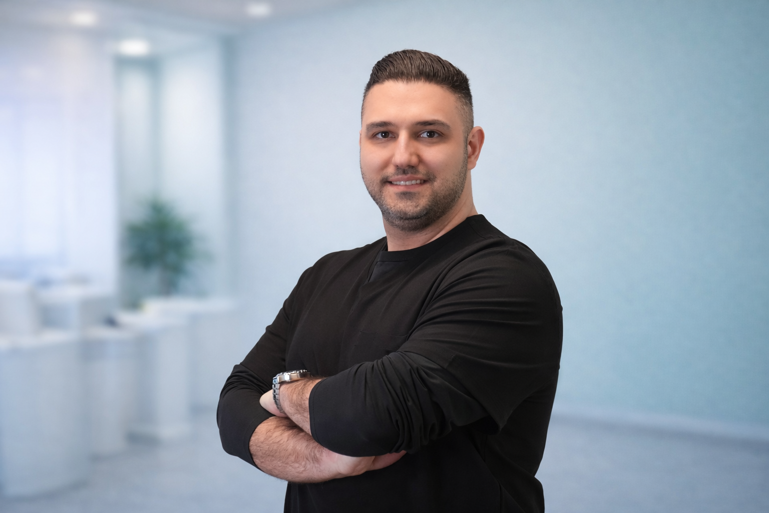 A man with short dark hair and a beard, wearing a black long-sleeve shirt, standing with arms crossed in a bright, modern office with a blurred background.