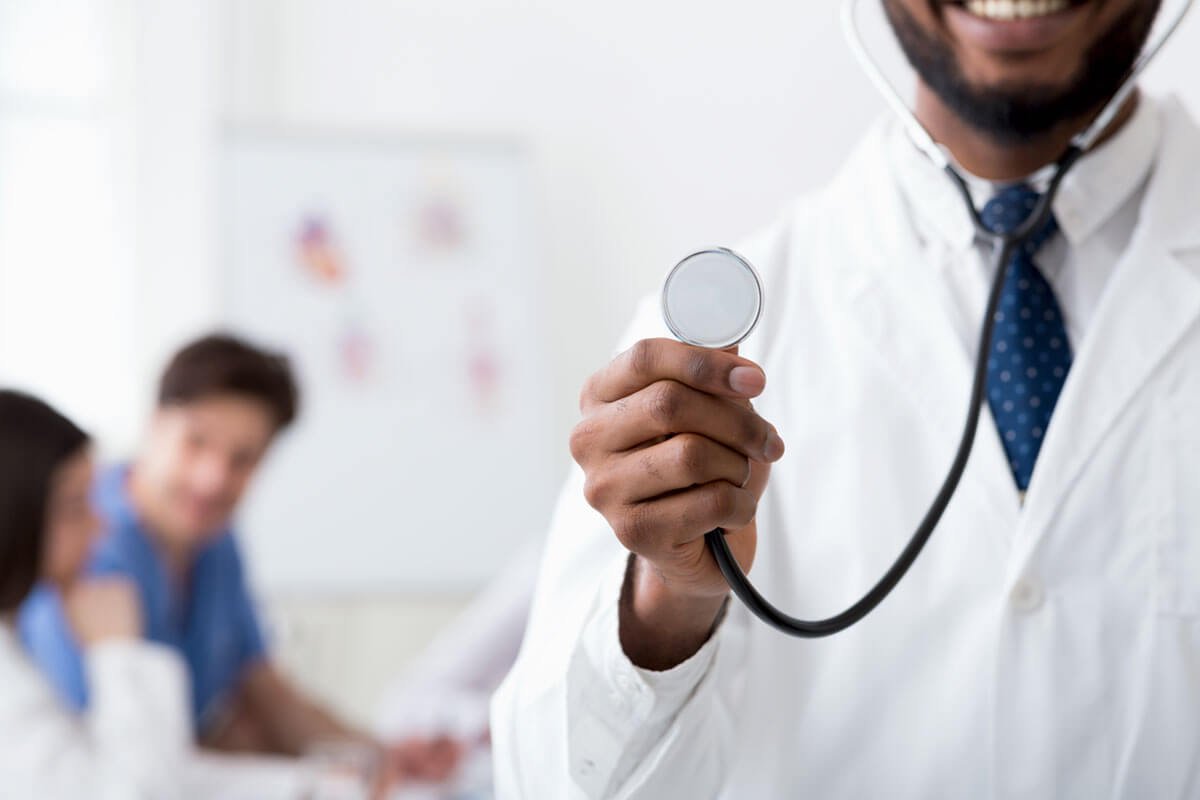 A male doctor in a white coat holding a stethoscope in a hospital or clinic, with two other medical professionals in the background.