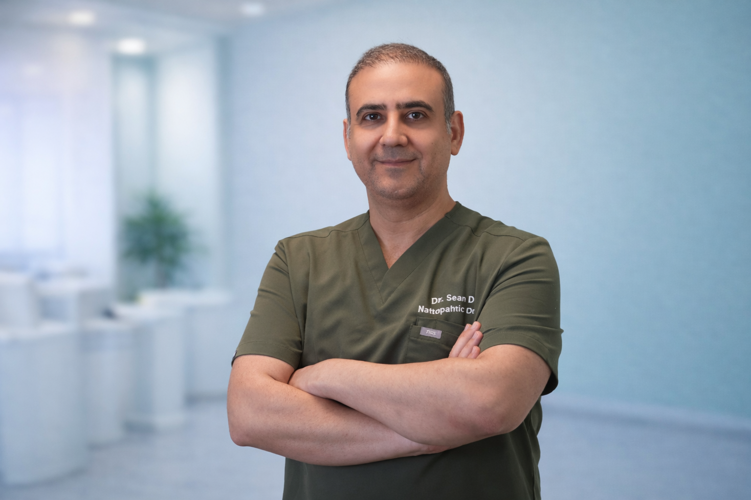 A male medical professional wearing green scrubs with his arms crossed, standing in a bright, modern clinic or hospital corridor.