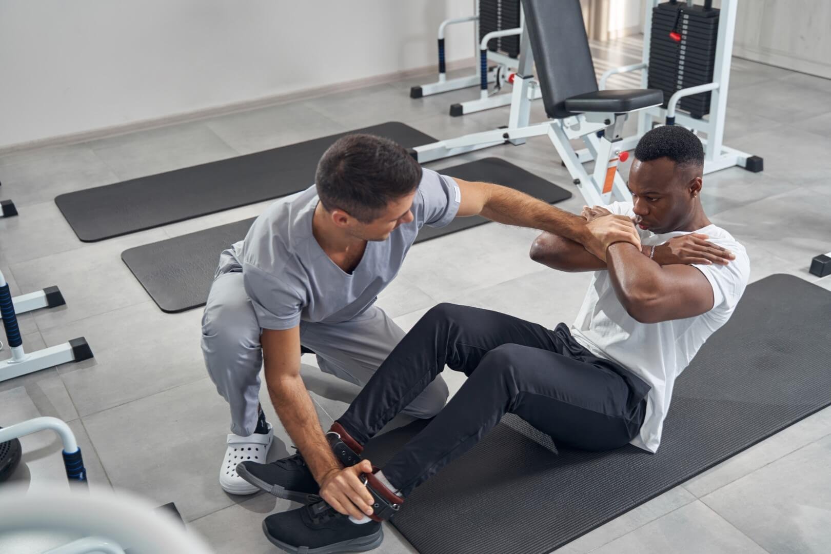 A personal trainer assisting an African American man in a white t-shirt with sit-up exercise in a gym.