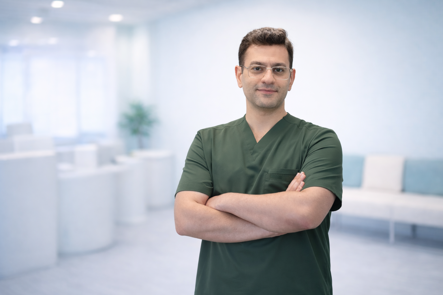 A male healthcare professional wearing green scrubs and glasses standing in a bright, modern medical office with arms crossed.