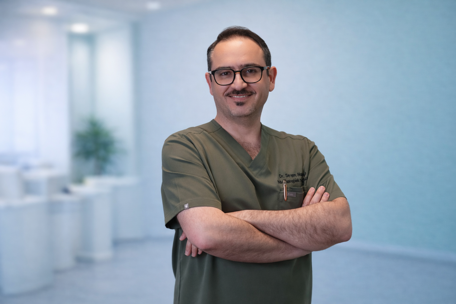 A male healthcare professional with glasses, wearing green scrubs, standing with his arms crossed and smiling in a bright, modern medical office or hospital corridor.