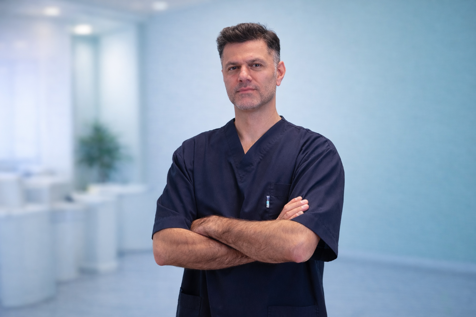 A middle-aged male doctor in navy scrubs standing with arms crossed in a bright, modern healthcare facility.