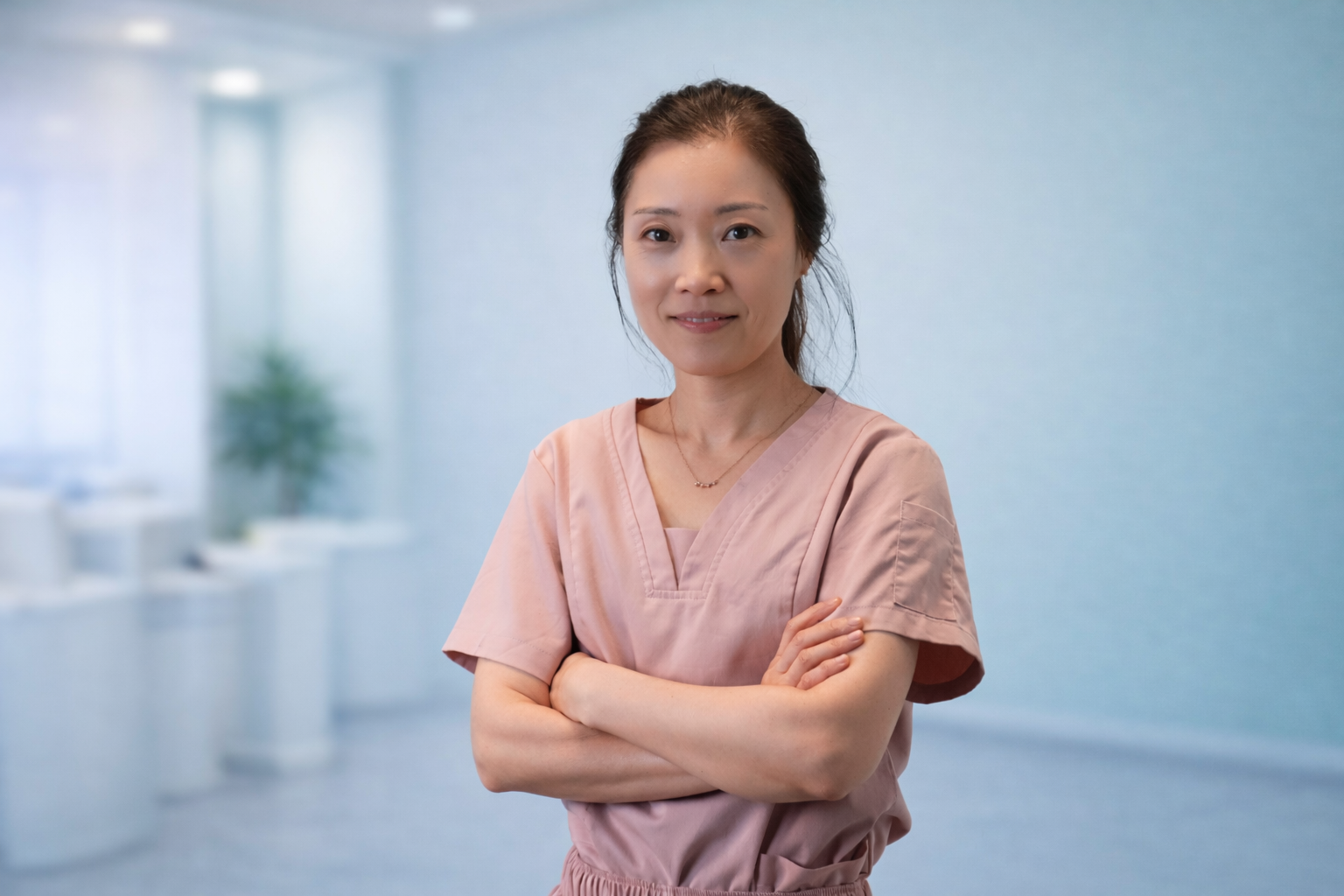 Portrait of a female healthcare professional in pink scrubs with arms crossed, standing in a medical facility with a blurred background.