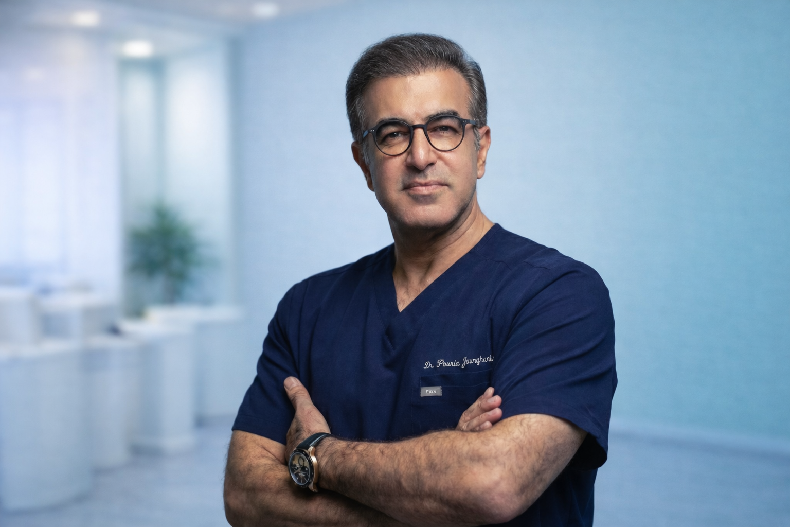 A confident male doctor in navy blue scrubs with glasses, arms crossed, standing in a modern medical clinic or hospital corridor with a blurred background.