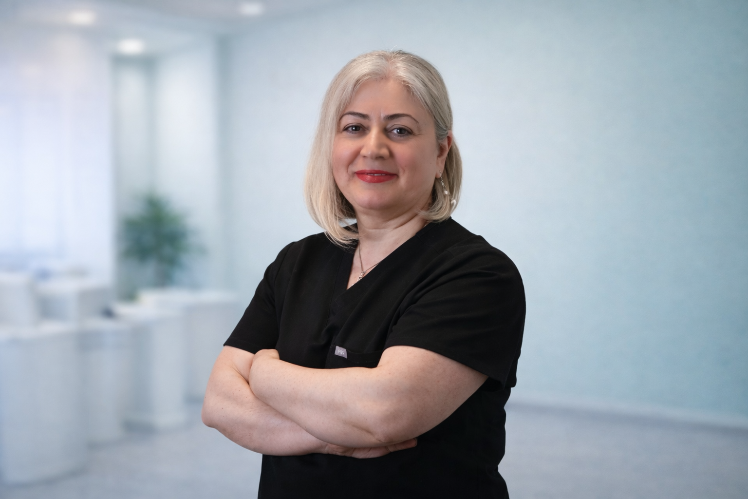 A confident woman with blonde hair, wearing black medical scrubs, standing in a modern, bright office with arms crossed and a slight smile.