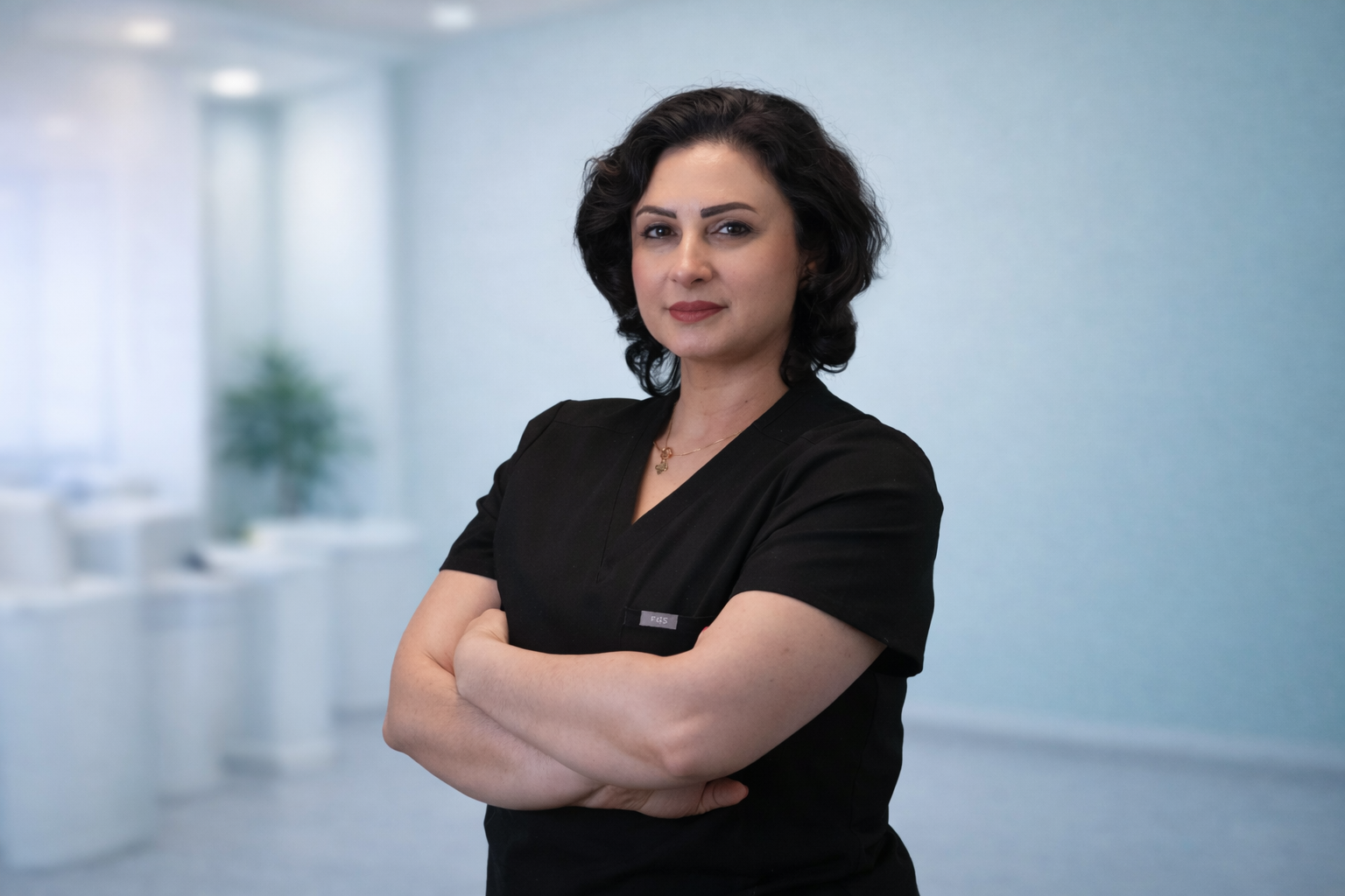 A woman with dark, curly hair wearing black scrubs, standing with arms crossed in a clinical setting.
