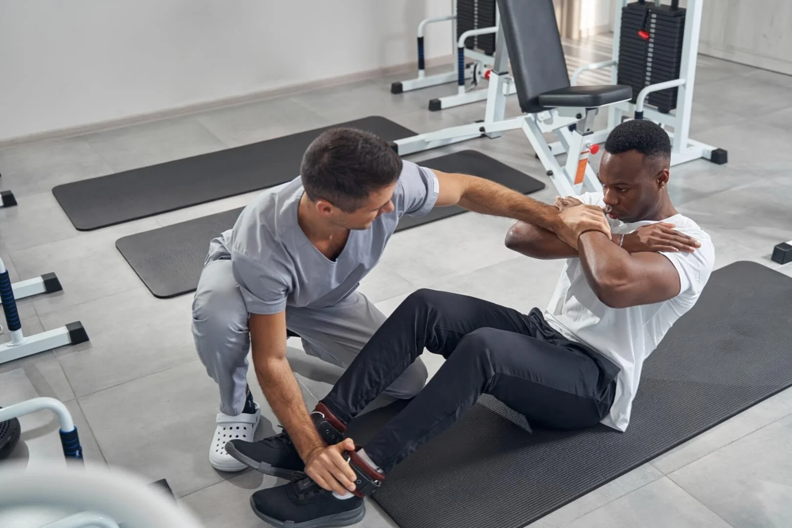 Physical therapist assisting a man with sit-up exercises on an exercise mat in a gym.