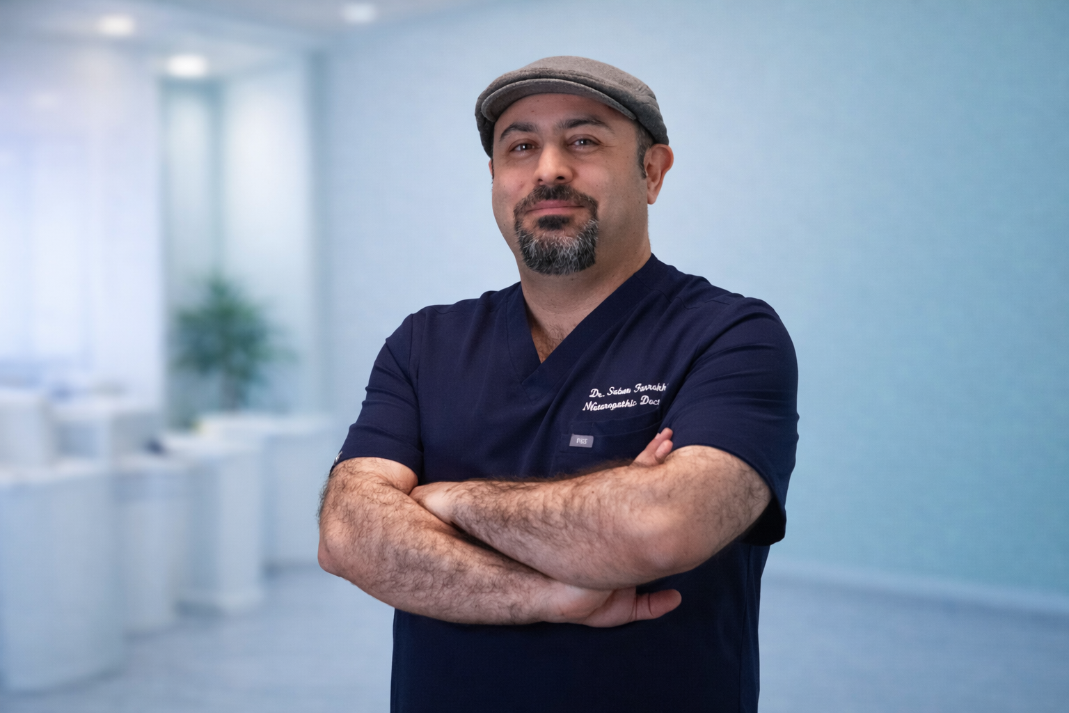 A man in a dark blue medical uniform with arms crossed, standing in a bright, modern, and clean healthcare or clinical setting. He is wearing a flat cap and has a beard and mustache, smiling slightly.