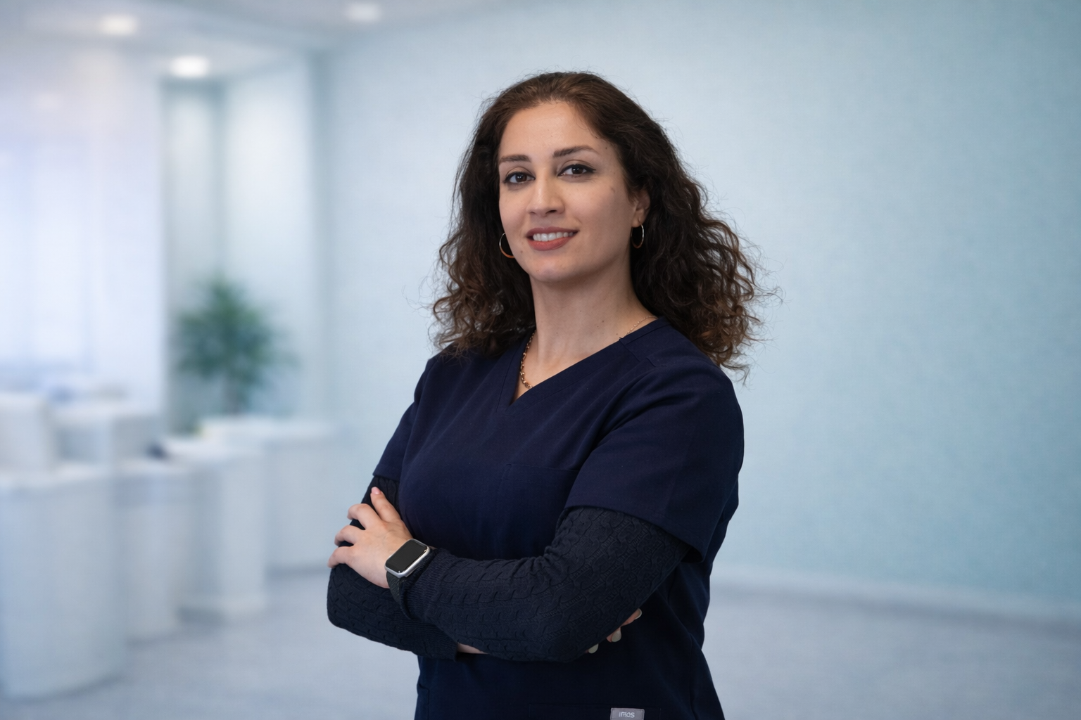 A professional woman with curly brown hair, wearing a dark blue scrub top, a smartwatch, and jewelry, standing with arms crossed in an office setting.