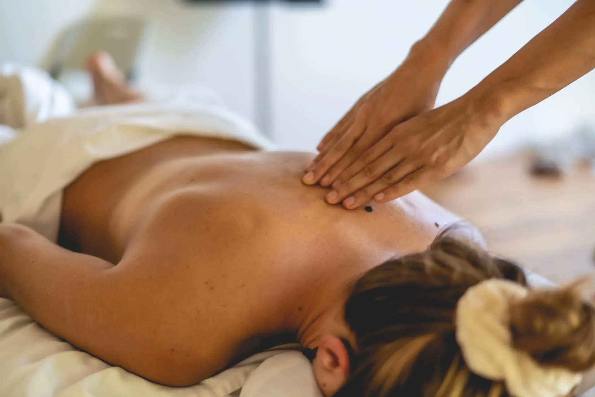 A person lying face down on a massage table receiving a back massage from a therapist. The person has long hair, and the therapist's hands are pressing into their back.