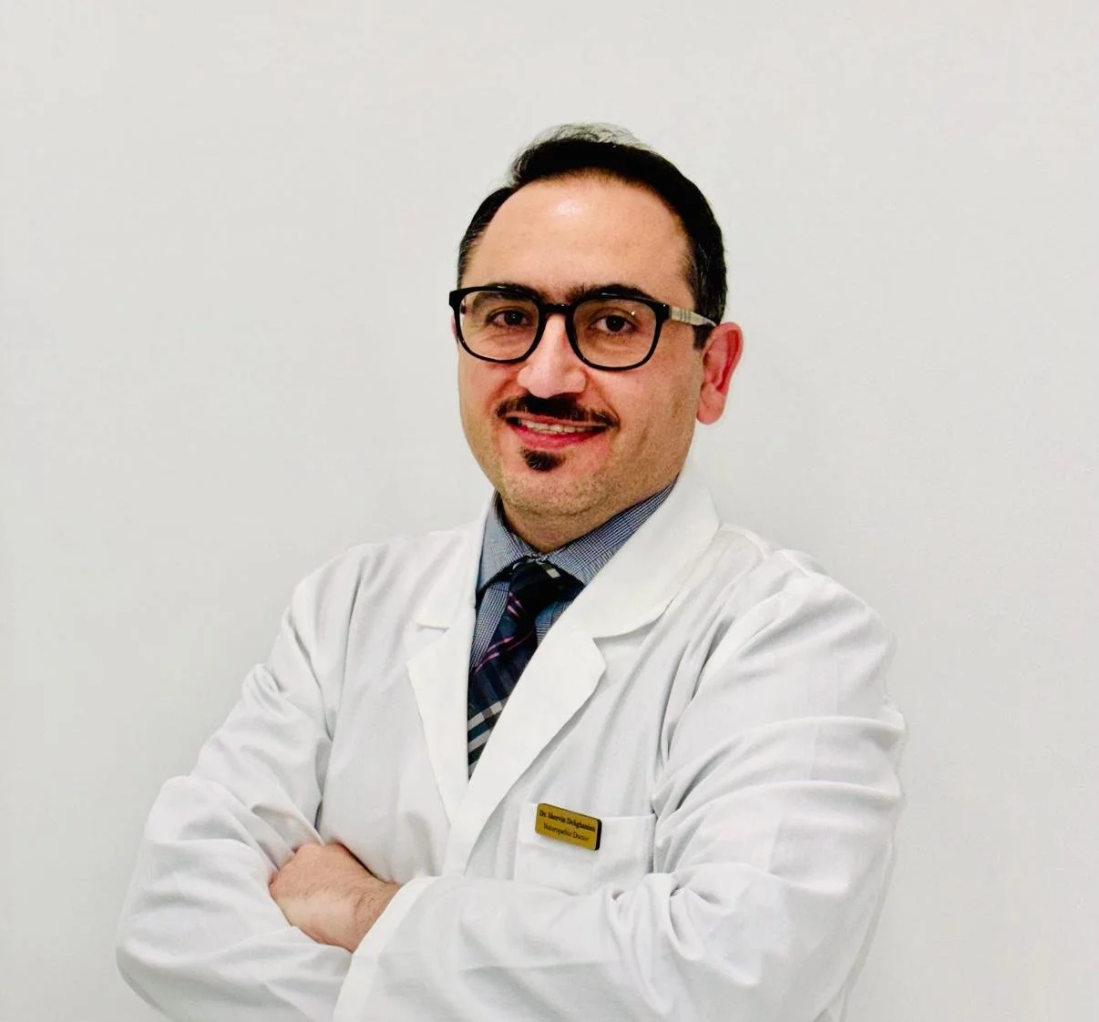 A male doctor with glasses, a mustache, and a goatee, wearing a white coat and patterned tie, standing with arms crossed in front of a plain white wall.