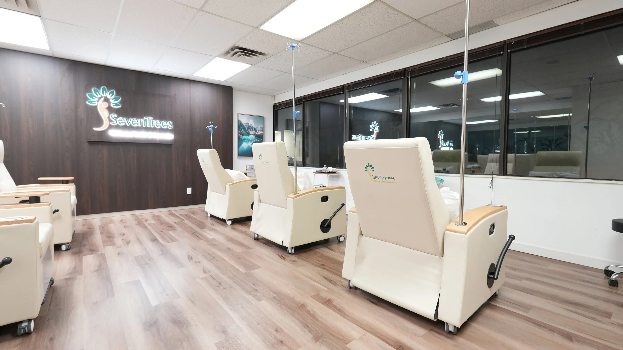 An empty hospital or medical treatment room with multiple reclining chairs, IV stands, and a dark wooden accent wall with a SevenTrees logo, large windows, and a landscape painting.