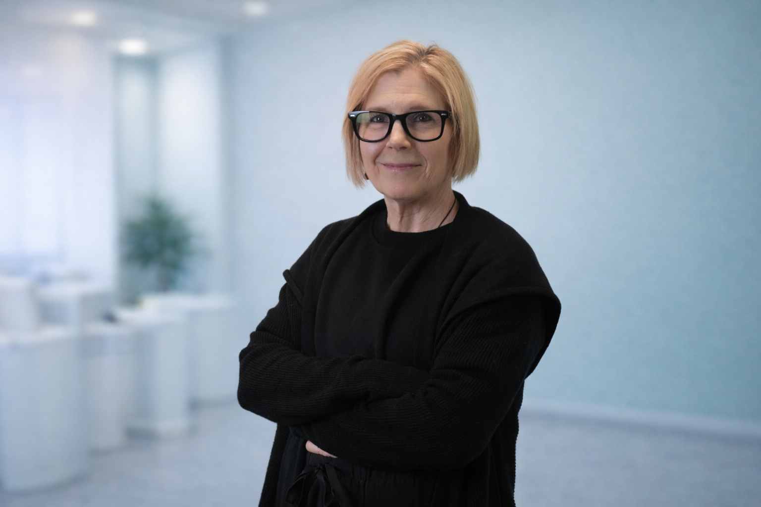 A woman with blonde hair, glasses, and a black outfit standing in a bright, modern office with a blurred background and potted plant.