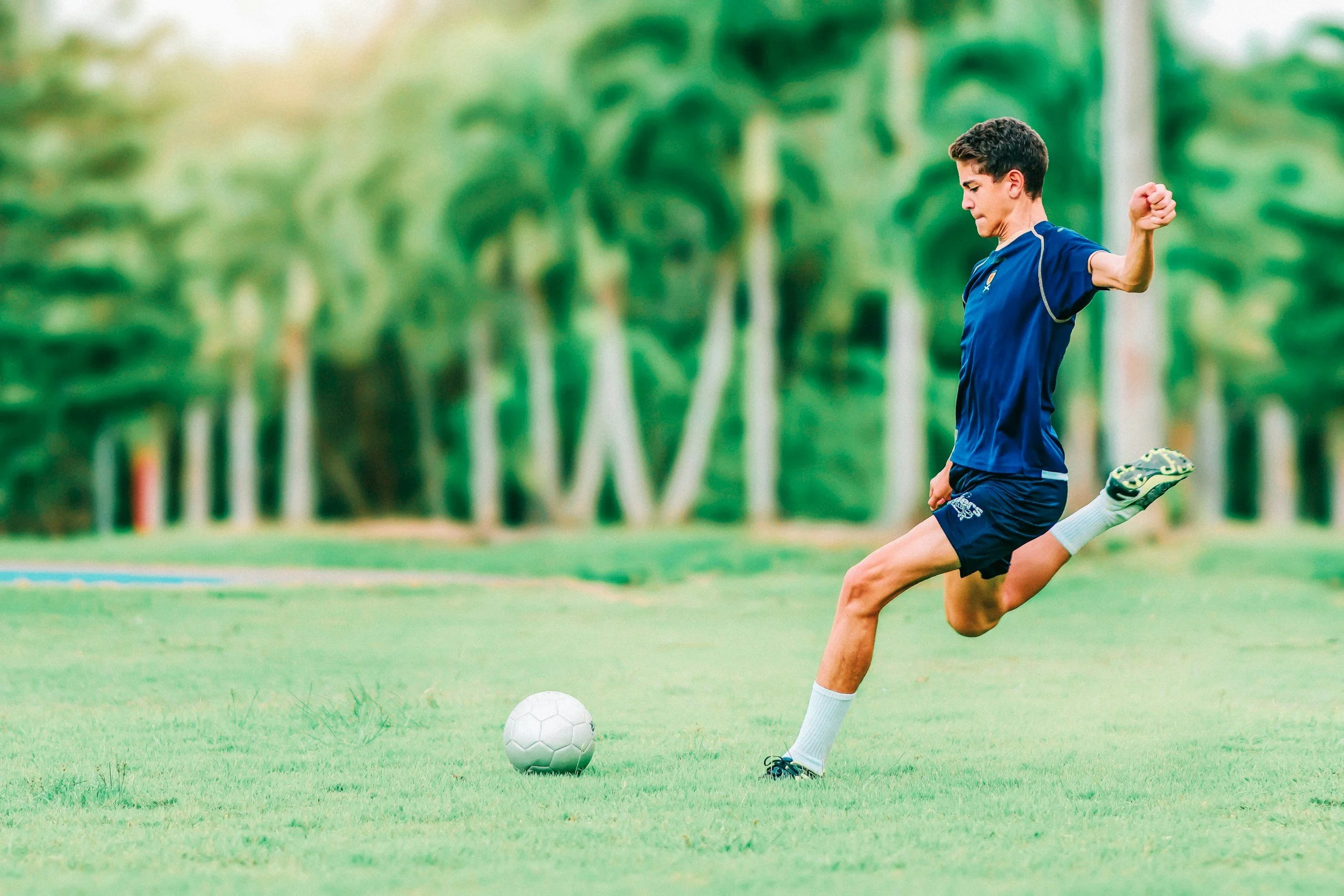 A young male soccer player in a blue uniform is kicking a white soccer ball on a grassy field with trees in the background.