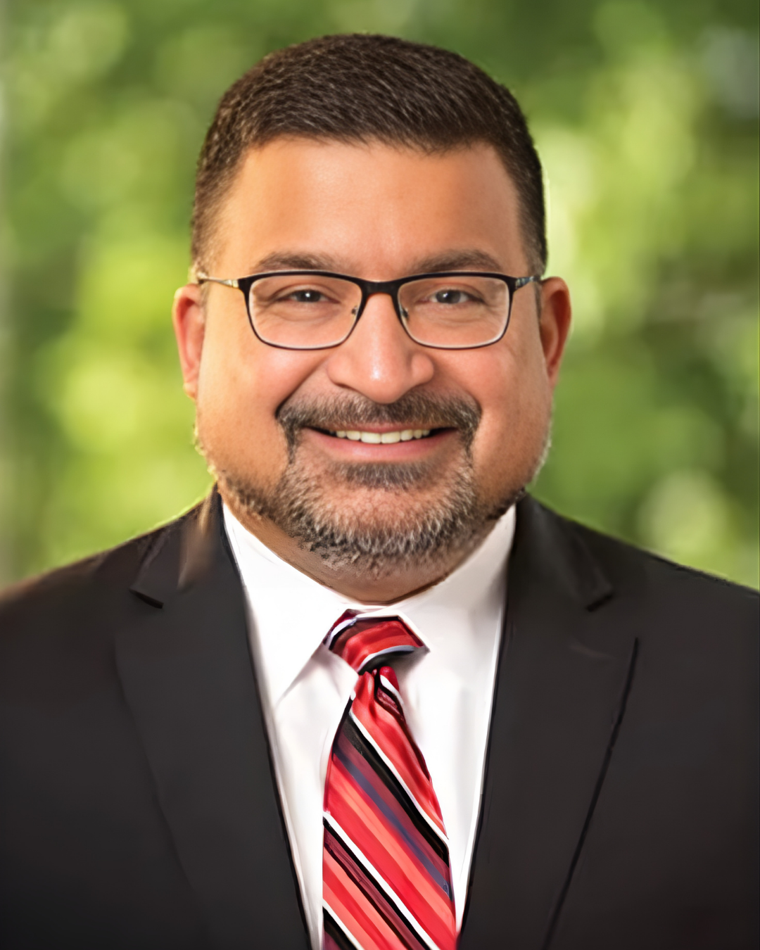 A man with glasses, a beard, and short dark hair, wearing a black suit, white shirt, and a red, black, and white striped tie, smiling in front of a blurred green background.