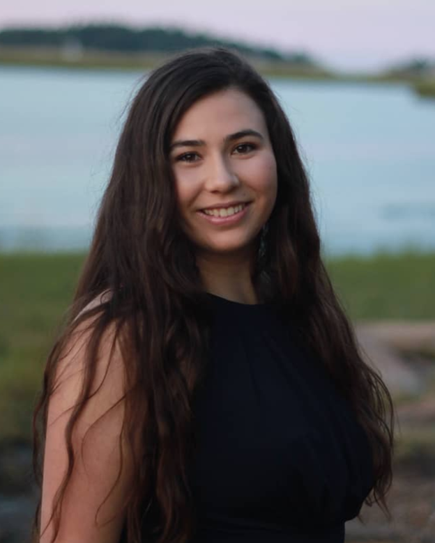 A young woman with long dark wavy hair smiling outdoors by a lake, wearing a sleeveless black top.