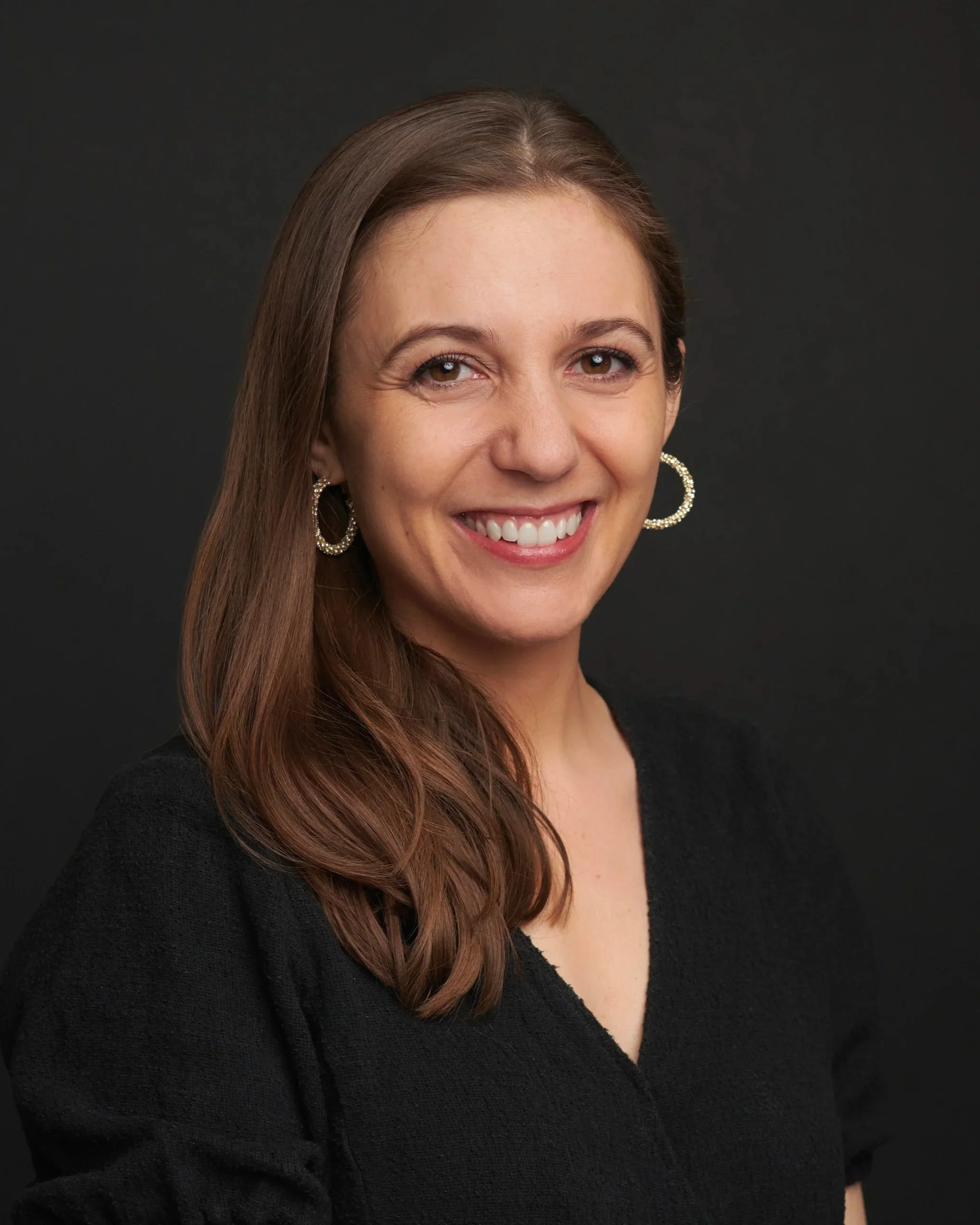 A woman with long brown hair smiling, wearing a black top and hoop earrings, against a dark background.