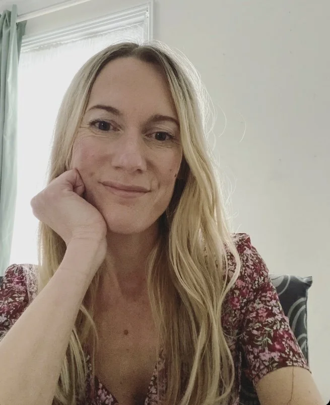 A woman with long blonde hair, smiling gently, resting her head on her hand, sitting indoors near a window with green curtains.