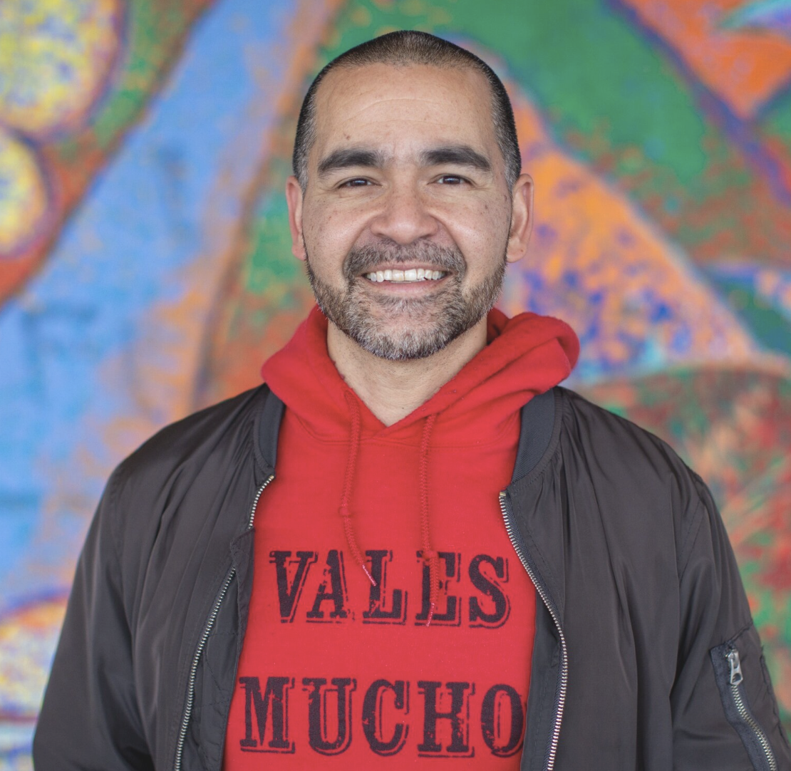 Man smiling, wearing a red hoodie with 'VALES MUCHO' text, against a colorful abstract background.