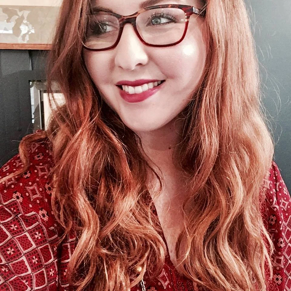 Smiling woman with red hair and glasses, wearing a patterned red top.