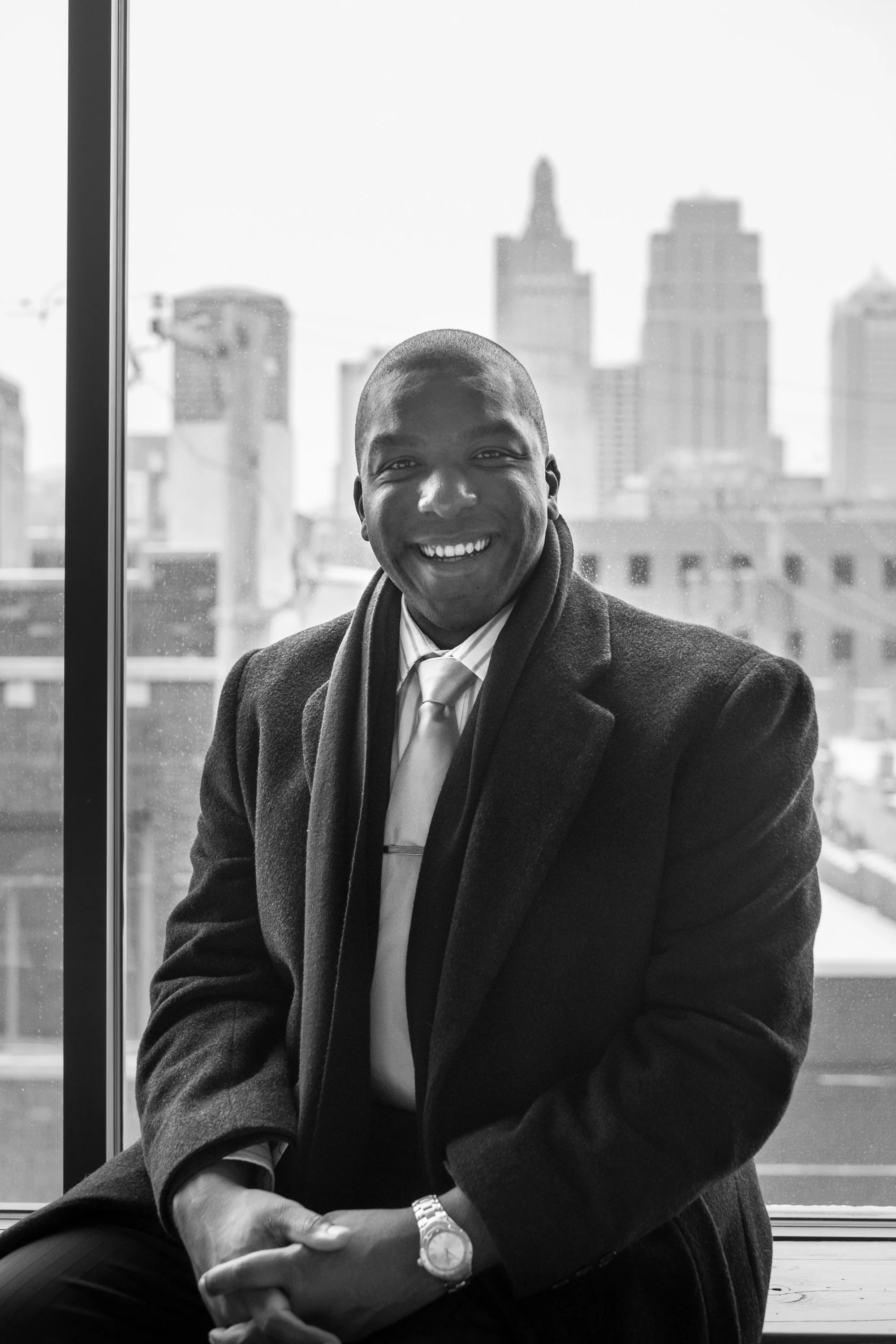 Smiling man in a coat and scarf sitting by a window with a cityscape background.