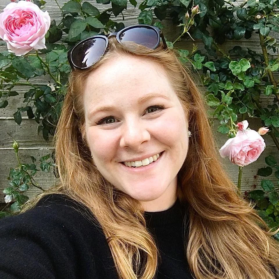 Woman smiling in front of pink roses and a wooden fence, wearing sunglasses on her head.