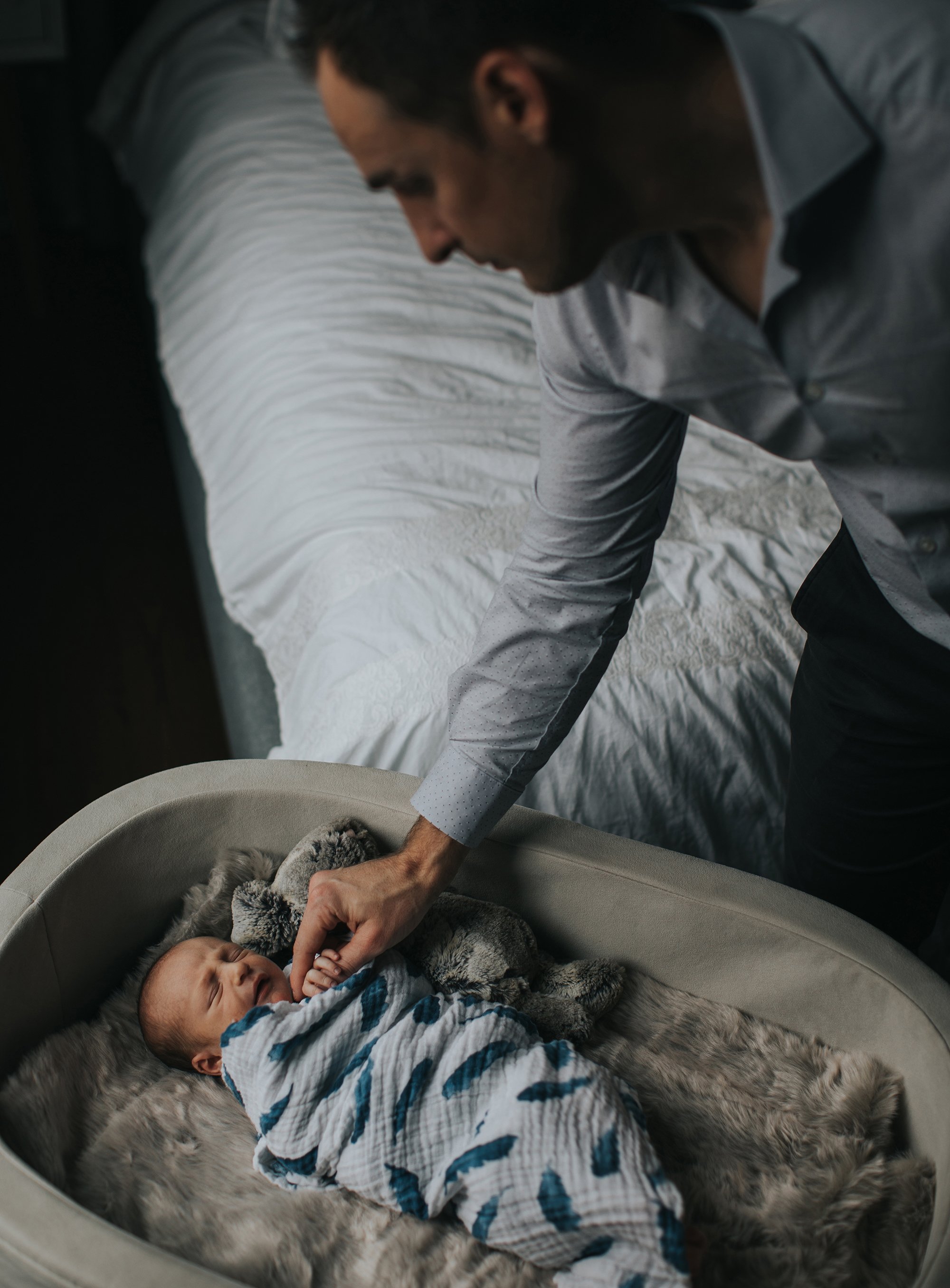 A father gently holding his newborn in a cozy cradle beside a bed, capturing a tender moment.