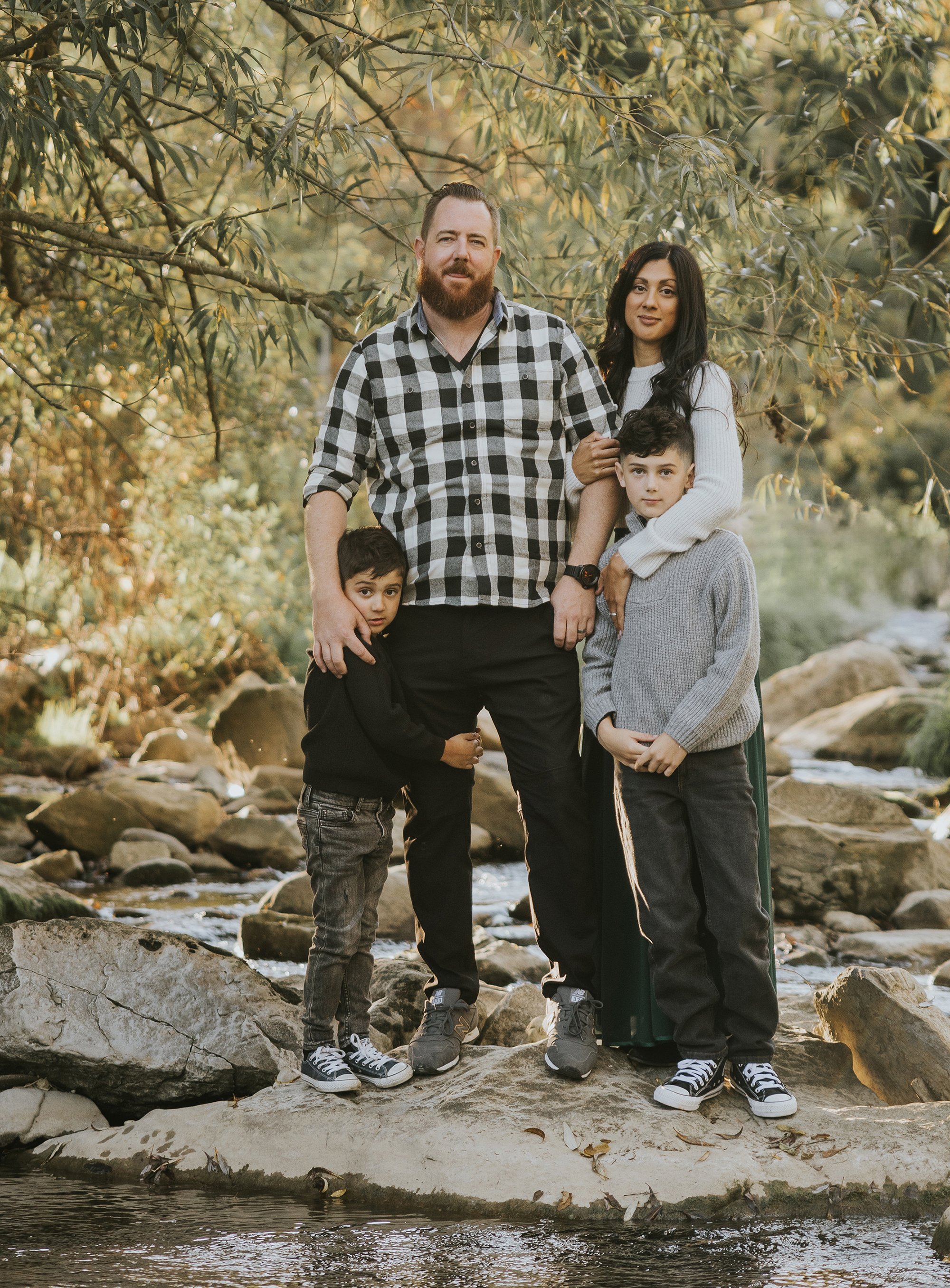 A family of four standing by a creek in a forested area, with natural light filtering through the trees, showcasing their connection in a serene outdoor setting.