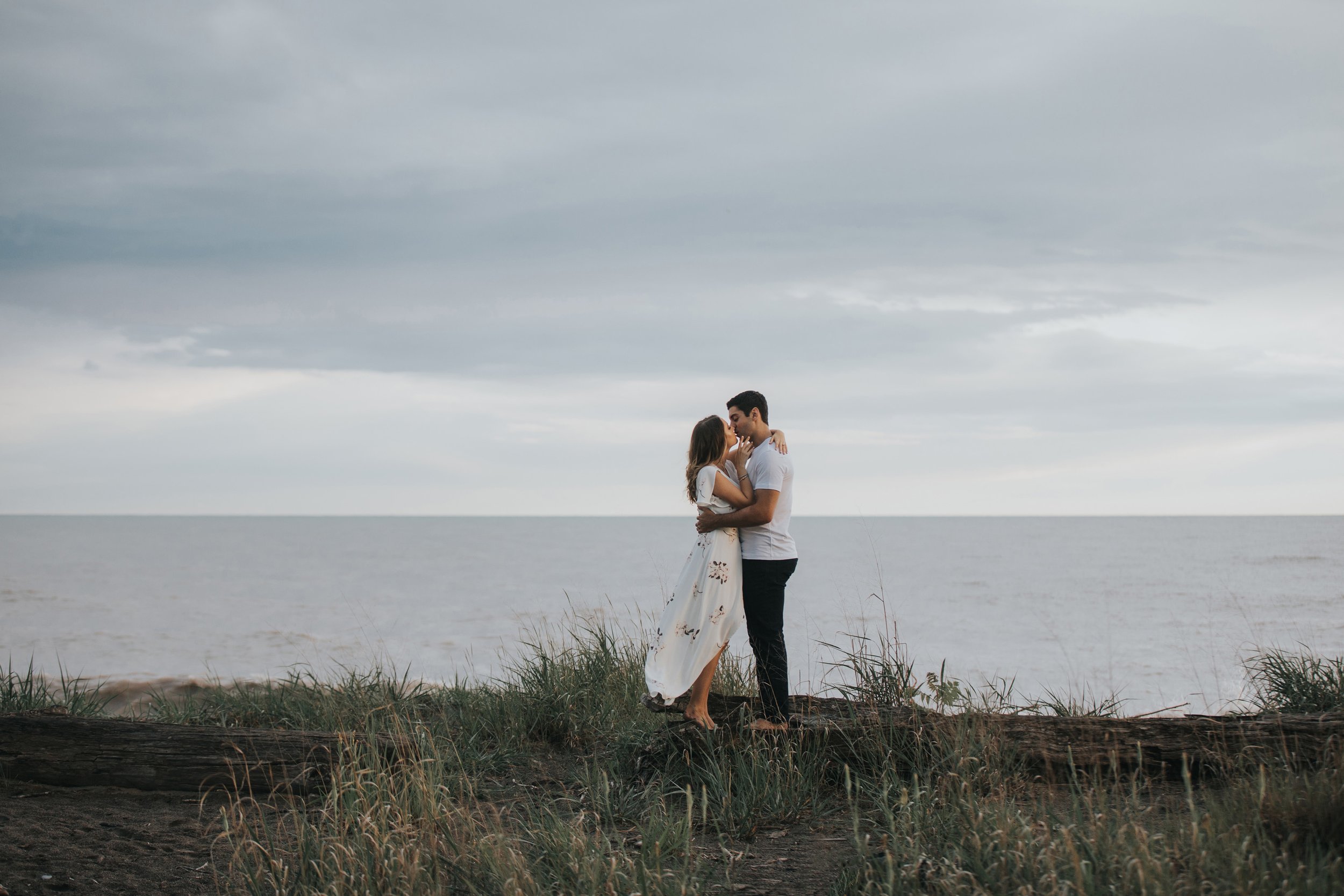 A couple shares an intimate embrace on a beach, with tall grass and the calm horizon in the background, creating a serene and romantic scene.